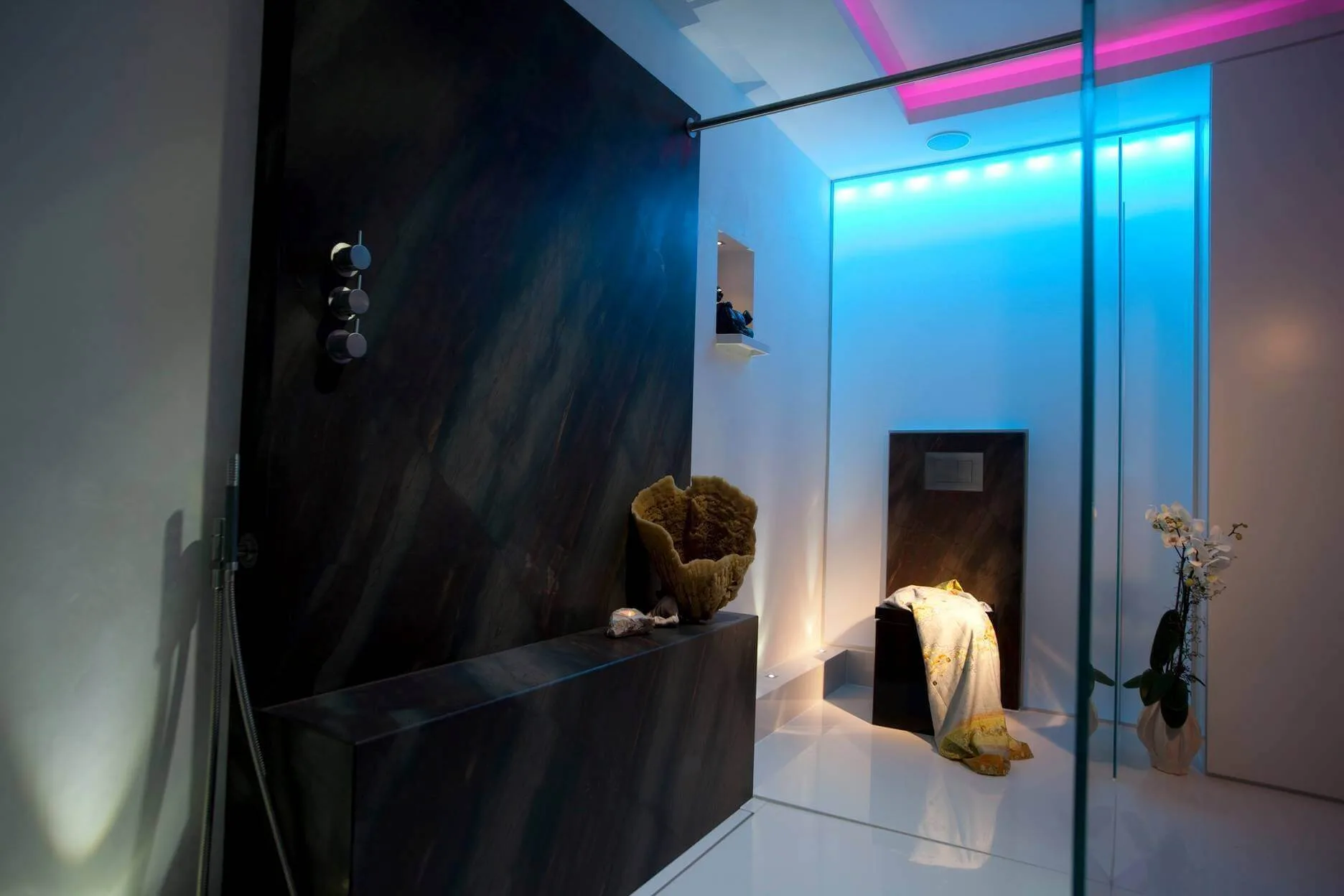 Die Bad Design Ideen vom Badplaner Torsten Müller umfassten für dieses Design Bad: Ein schwarzes WC als exklusiven Akzent im Bad eine beheizte Sitzbank in der Dusche zum barrierefreien Duschen Accessoires von Vola Edelstahl gebürstet für einen modernen Eindruck indirekte Beleuchtung durch Lichtvoute und LED RGB hinter einer Wand- und Deckenbespannung schlichtes Baddesign in der Detailplanung, um ein meditatives Badambiente zu erzeugen. Die natürliche Leidenschaft für Stein kommt auch beim Badezimmer planen in dieser Badgestaltung durch die Verwendung des Naturmaterials von Antolini Verona, das auch in der Badezimmerausstellung der Bäder München zu sehen ist, zum Ausdruck. Ein harmonisches Badambiente im Design Bad entstand beim Badezimmer Gestalten durch das Bad Planen mit dem Antolini Naturstein, dessen Bad Muster für beste Badezimmer in der Bäderausstellung in Bonn, Köln, Bad Honnef oder Düsseldorf vorhanden sind. Anstatt auf eine Design Badewanne fiel die Wahl bei der Badezimmer Design Badgestaltung auf eine Dusche, die Golden Ager sicheres und barrierefreies Duschen ermöglicht. Die Dusch-Sitzbank aus braunem Naturstein aus der Bäderausstellung der Antolini Stone Gallery ist beheizt. Der großzügige Dusch-Bereich im Designer Bad wurde mit 13 Millimeter starken Sicherheitsglas abgetrennt. Eine moderne Vola Kopfbrause verspricht entspannte Momente im Komplettbad unter dem vitalisierenden Wasserstrahl. Best Ager Bad Design