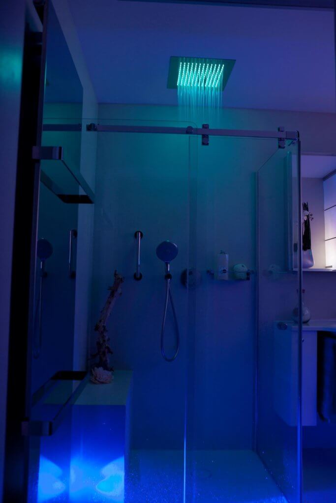 Stimmungsvolle Lichtszenarien im Badezimmer Die Farbtherapie harmonisiert das körperliche und das seelische Wohlbefinden mit Hilfe von Farben. Diese verfügen über eine bestimmte Wellenlänge, die je nach Farbton einen entspannenden oder vitalisierenden Effekt aufweist. Die hinter einer transparenten Glaswand verborgene Regendusche mit LED Leuchten besitzt eine intuitiv zu bedienende Schaltfläche, auf der die einzelnen Impulse individuell eingestellt werden können. Auch eine Programmierung im Voraus erlaubt es in der Früh mit den Strahlen einer aufgehenden Sonne aufzustehen. In dem vorliegenden Projekt wurde der Colorshower der Firma KWS eingebaut, der über moderne Lichtelemente in der Kopfbrause verfügt. Vielseitige Funktionen, wie „Wave“, „Sweep“ und viele mehr lassen die LEDs in einem vorgegebenen Rhythmus aufleuchten, der je nach Wunsch beruhigt oder aufweckt. Auf der beheizten Sitzbank in der bodeneben gestalteten Dusche kann man sich eine Weile von dem Alltag entspannen und all den Stress hinter sich lassen, während von oben warme Wasserstrahlen herabfallen.