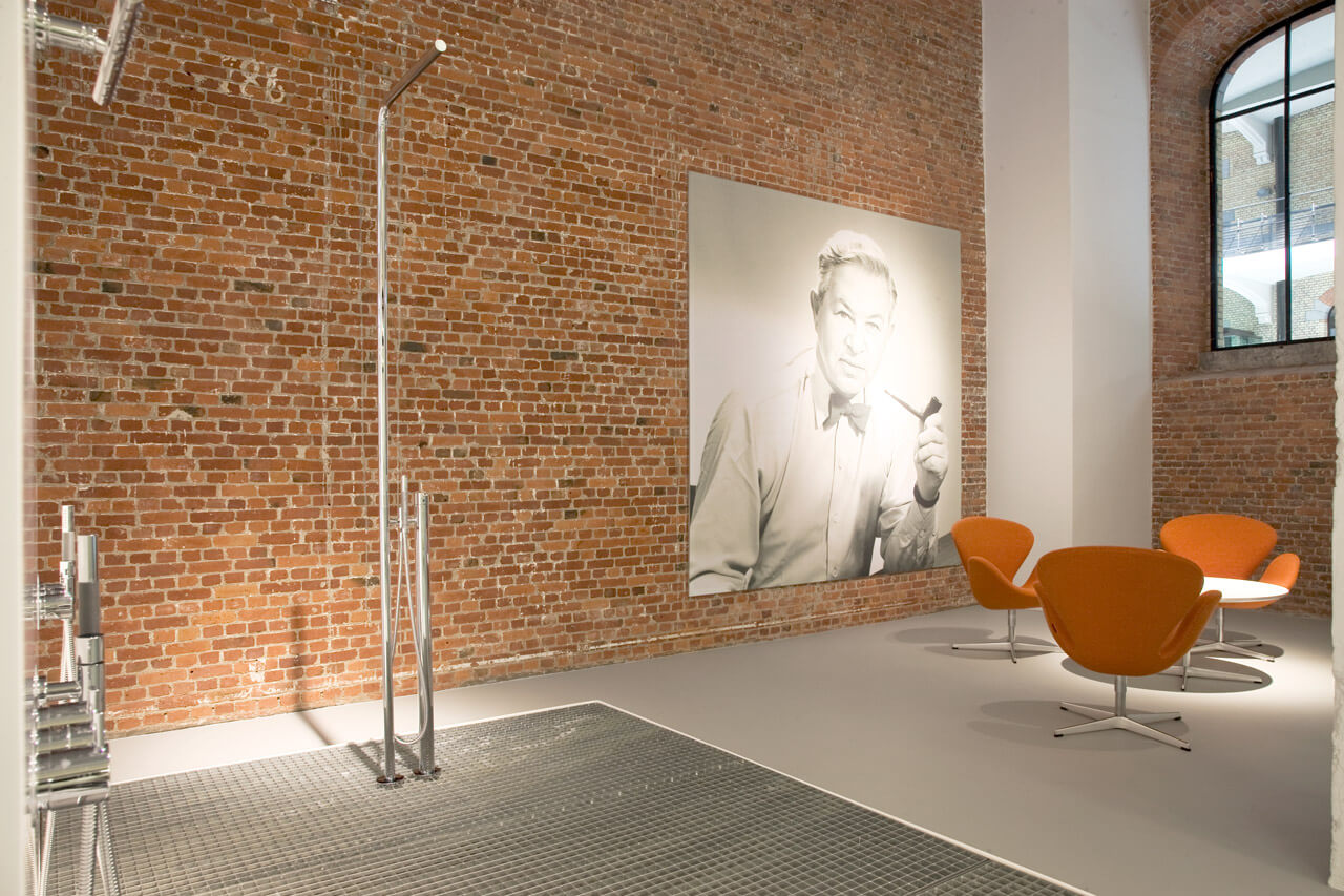 Präsentation von Vola Duscharmaturen vor einer Ziegelwand mit Foto von Arne Jacobsen und Sitzbereich