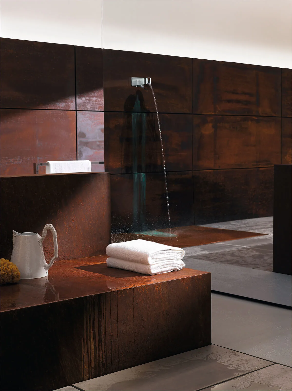 Elemental Spa Die Materialien Cortenstahl: Cortenstahl, auch CORTEN-Stahl genannt, zeichnet sich durch eine Oberfläche aus, die mit einer besonders dichten Eisenoxidschicht überzogen ist. Hierdurch verleiht Feuchtigkeit dem Material eine besonders individuelle, ausdrucksvolle Patina. Gleichzeitig schützt die Schicht den Stahl vor Verwitterung. Daher wird Cortenstahl auch zur Akzentuierung in der Architektur und als Werkstoff in der Kunst eingesetzt. Wird das Material – wie in einigen Bereichen des ELEMENTAL SPA – zusätzlich mit einer speziellen Kupferlegierung versehen, lassen sich die Spuren des Wassers noch starker betonen. Es entsteht durch den Gebrauch eine Topografie der individuellen Reinigungsrituale – elementar und modern. Kupfer: Kupfer ist ein warmes Metall mit verschiedenen Eigenschaften und einer besonderen Ausstrahlung. In Verbindung mit Wasser entsteht Grünspanbildung und damit eine sehr eigene optische Anmutung. Als Wannenmaterial wird Kupfer darüber hinaus ein therapeutischer Effekt zugesprochen. Marmor: Ein edles Material, das trotzdem Ursprünglichkeit vermittelt. Massiv aus dem Schoß eines Berges geschlagen. Durch seine Schwere trägt Marmor zu einem Gefühl der Entschleunigung bei. Corian® : Überzeugt durch seine Materialität: ein stabiler und wasserunempfindlicher Werkstoff aus acryl-gebundenem Mineral, der unendlich viele Designmöglichkeiten mit extremer Langlebigkeit verbindet. Olivenholz: Die Möbel im ELEMENTAL SPA sind aus Olivenholz in unterschiedlichen Tönen gefertigt. Das langsam gewachsene Hartholz zeichnet sich durch besonders dichte, unempfindliche Poren aus. Olivenholz verbreitet eine natürliche Wärme und hat darüber hinaus in vielen Kulturkreisen eine spirituelle Bedeutung. Während diese Materialien allesamt eine Hommage an das Archaische und Ursprüngliche sind, lassen sich über die Dekoration gezielt Kontrapunkte setzen. Hochwertige Gegenstände, die das Lineare, Schlichte und Natürliche der Badarchitektur und der Materialien aufgreifen und dezenten Luxus ausstrahlen.