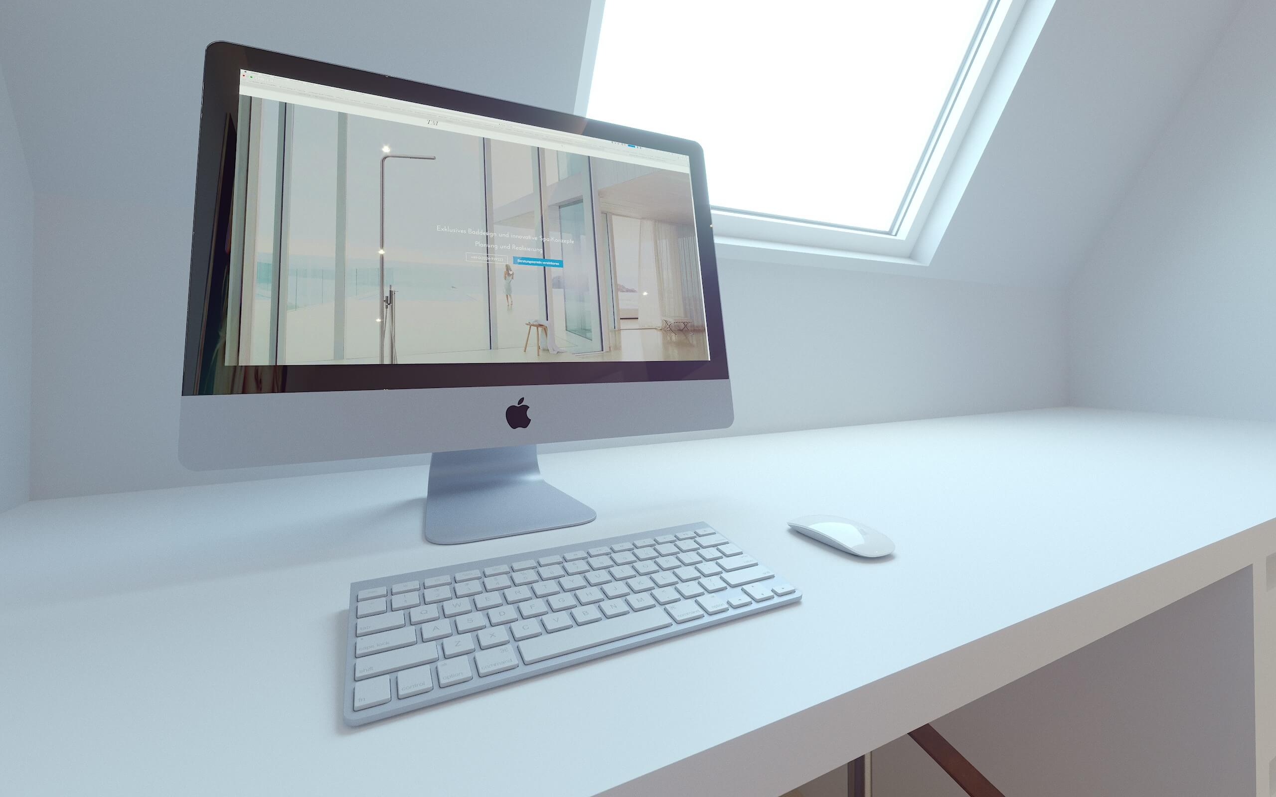 Apple-Monitor und weiße Tastatur auf weißem Schreibtisch vor einer Dachschräge mit Fenster