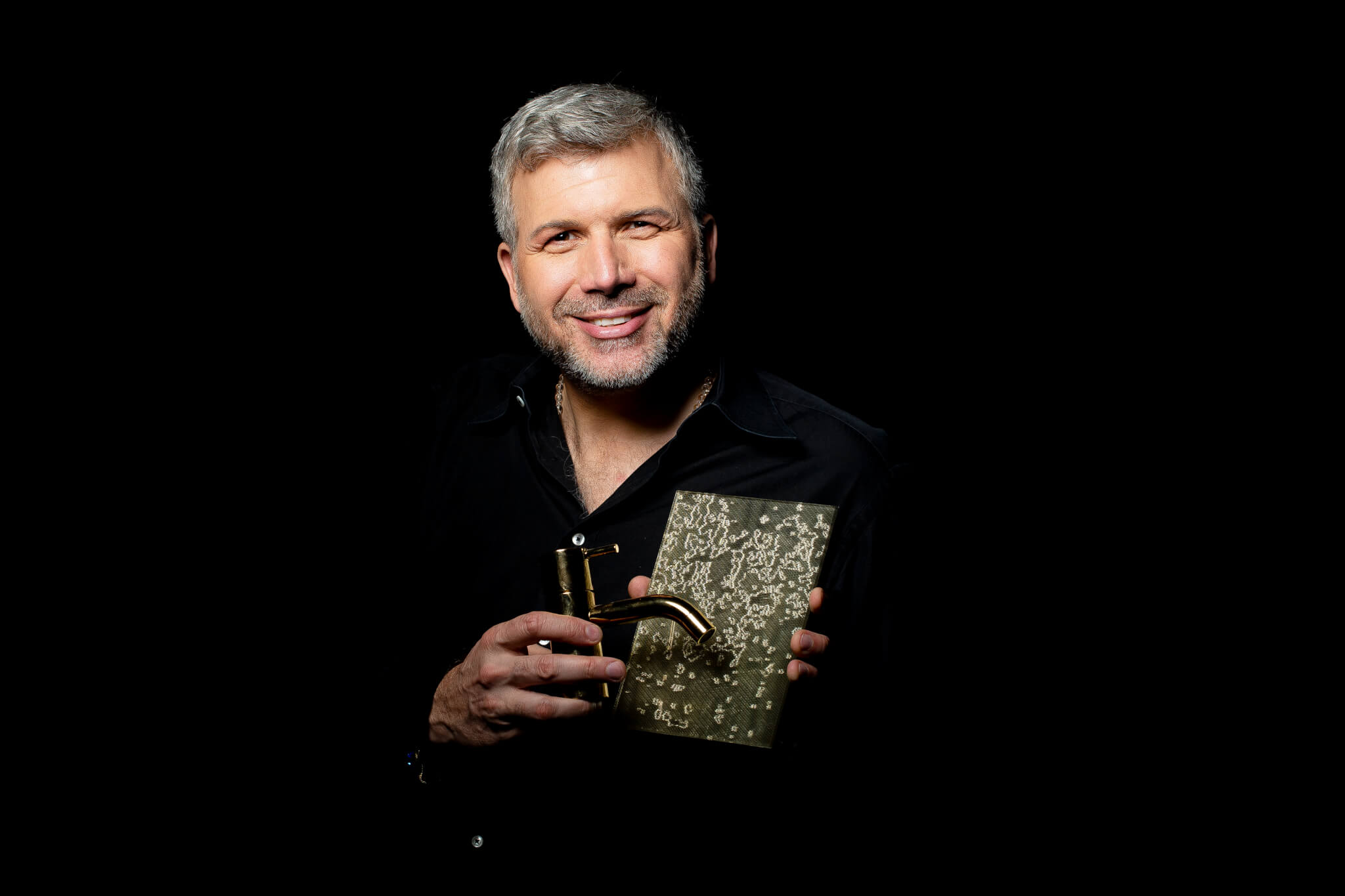 Torsten Müller hält lächelnd goldene Badezimmer Armaturen in den Händen.