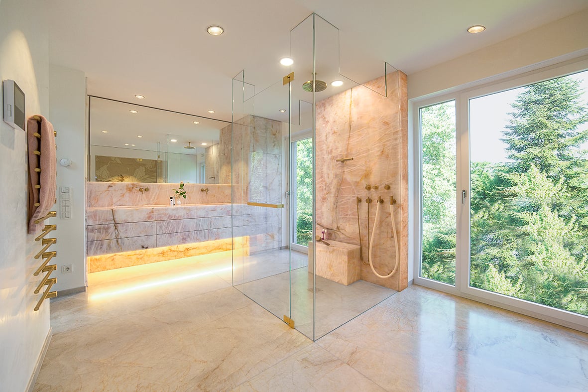 Das exklusive Badezimmerdesign wurde um einen rosa-pinkfarbenen Naturstein von Antolini aus Verona konzipiert. Dieser edle Naturstein verleiht dem Raum eine einzigartige Eleganz. "Cristallo Rosa WOW" gehört zu den ausdrucksstärksten Natursteinen innerhalb der exklusiven Kollektion von Antolini®. Die rosa Töne erinnern an die Essenz blühender Kirschbäume und entführen den Betrachter in eine erfrischende und ruhige Frühlingsoase. Helle, feine Linien ziehen sich über die gesamte Oberfläche und schaffen Dynamik in den Texturen, während dunklere Adern das Landschaftsbild zwischen Abstufungen von blassrosa durchziehen. Nur wenige Steine können eine so entspannende und wunderbare Wirkung erzeugen wie der Cristallo Rosa WOW. Die Designarmatur stammt von Arne Jacobsen, bekannt für seine Bauhaus-inspirierten Klassiker, und ergänzt das luxuriöse Ambiente perfekt. Die Badezimmergestaltung umfasst eine geräumige Dusche mit Kneippschlauch für kalte Güsse, Kopfbrause und Handbrause. Eine Badewanne wurde bewusst nicht integriert. Stattdessen bietet die große Dusche mit Schwallbrause ein erfrischendes Duscherlebnis. Das Neo-Rest-WC von Toto setzt Maßstäbe in Exklusivität, Technologie und Reinigungsfreundlichkeit. Die Waschtischanlage ist als Doppelwaschtisch mit großem Spiegel konzipiert. Unter dem eleganten Naturstein verbergen sich geschickt integrierte Schubladeneinheiten sowie die Waschmaschine und der Trockner. Diese Geräte sind hinter dem Naturstein versteckt, was zu einem nahtlosen Erscheinungsbild beiträgt. Eine Sauna von Klafs fügt sich harmonisch in das Gesamtdesign ein, während ein Apothekerauszugschrank zusätzlichen Stauraum bietet.