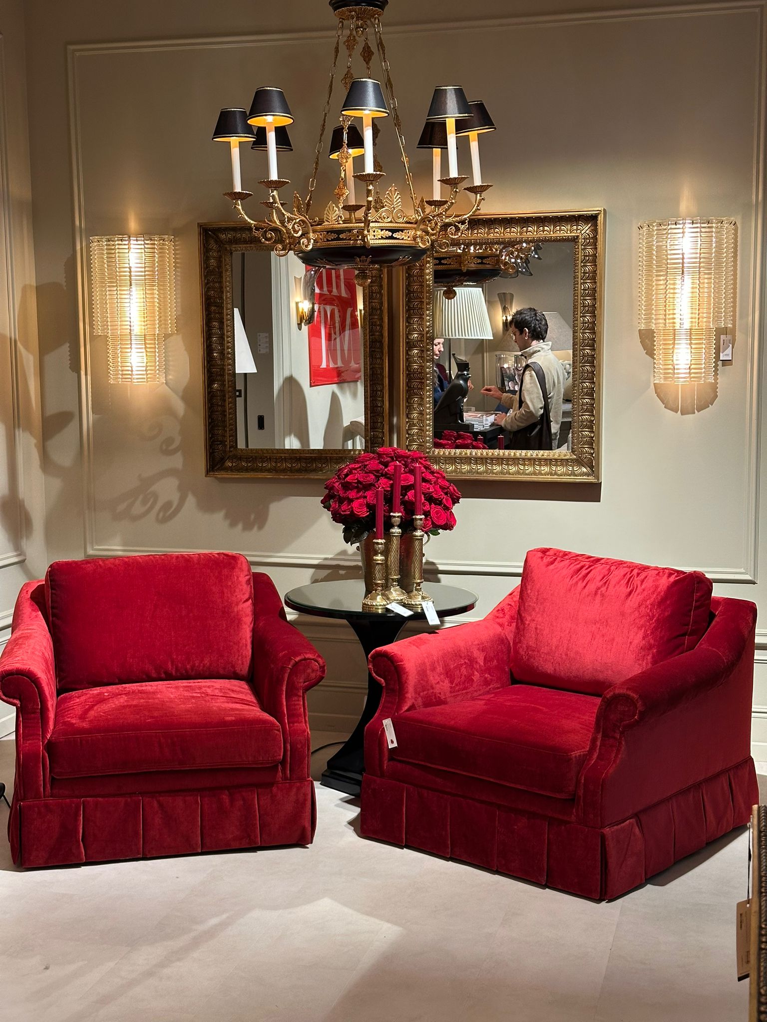 Zwei luxuriöse rote Samtsessel mit klassischem Design, flankiert von einem runden Beistelltisch und goldenen Akzenten. Im Hintergrund ein prunkvoller Kronleuchter und ein Spiegel auf der Maison & Objet 2025.