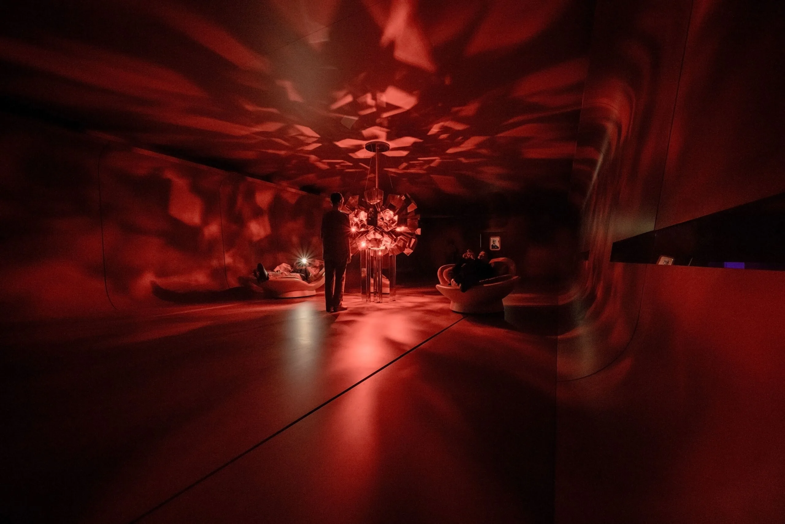 Künstlerische Lichtinstallation in tiefem Rot auf dem Salone del Mobile 2025 mit Besuchern in loungiger Atmosphäre.