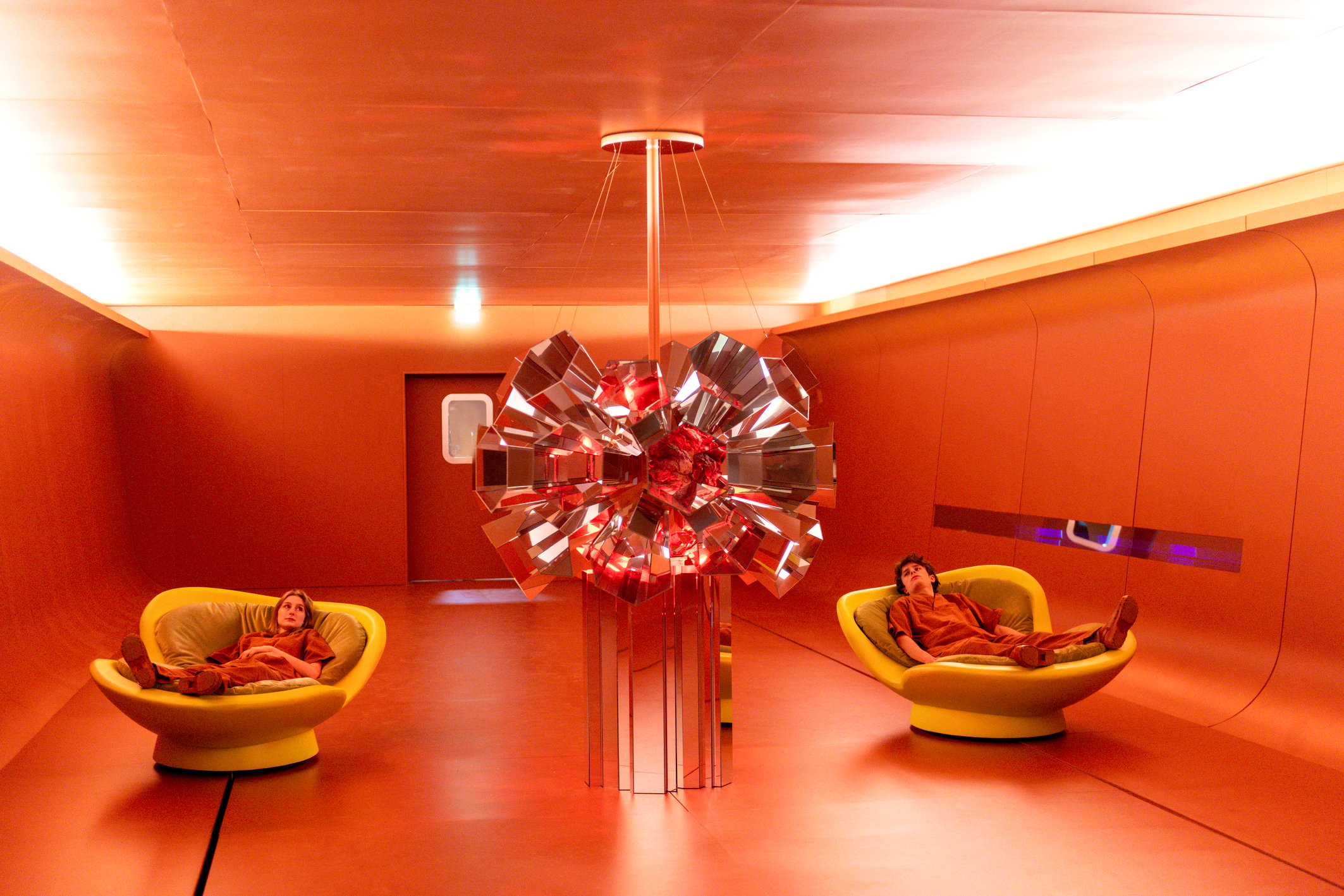 Zwei Personen entspannen in futuristischer, rot getauchter Rauminstallation mit zentralem Lichtobjekt auf dem Salone del Mobile 2025.