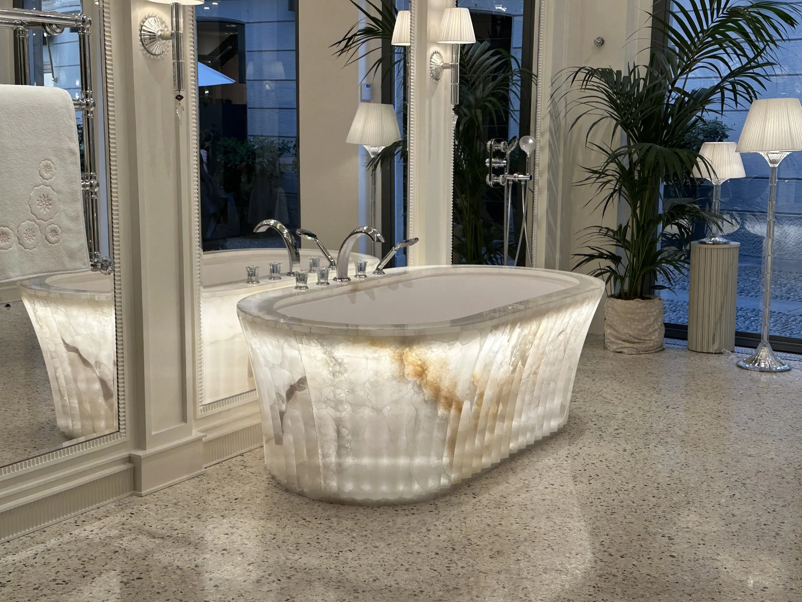 Beleuchtete freistehende Luxus-Badewanne aus Onyx in einem eleganten Spa-Badezimmer mit Spiegelflächen und Palmen.