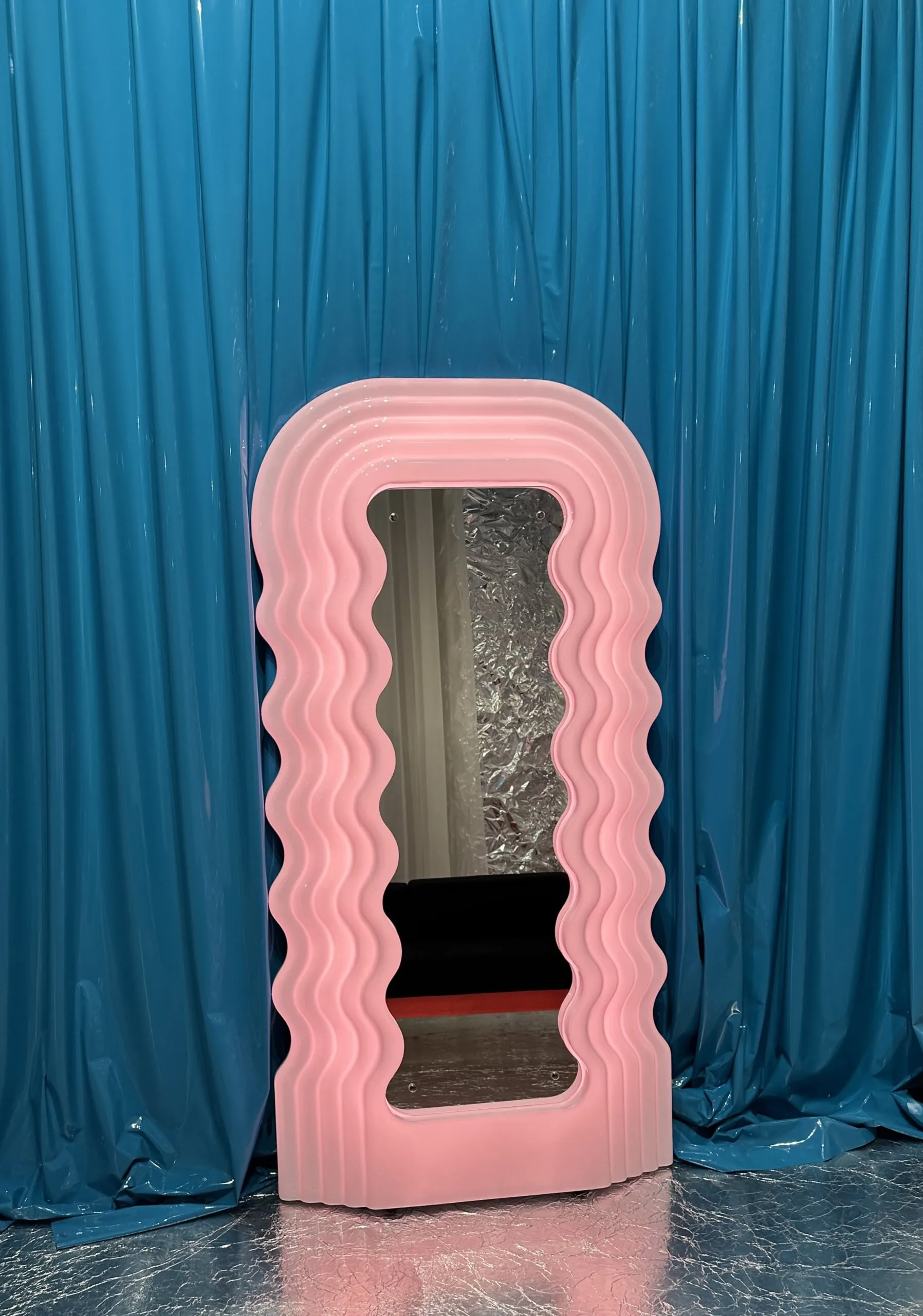 Welliger, pinkfarbener Designspiegel vor türkisfarbenem Vorhang auf dem Salone del Mobile 2025 in Mailand.