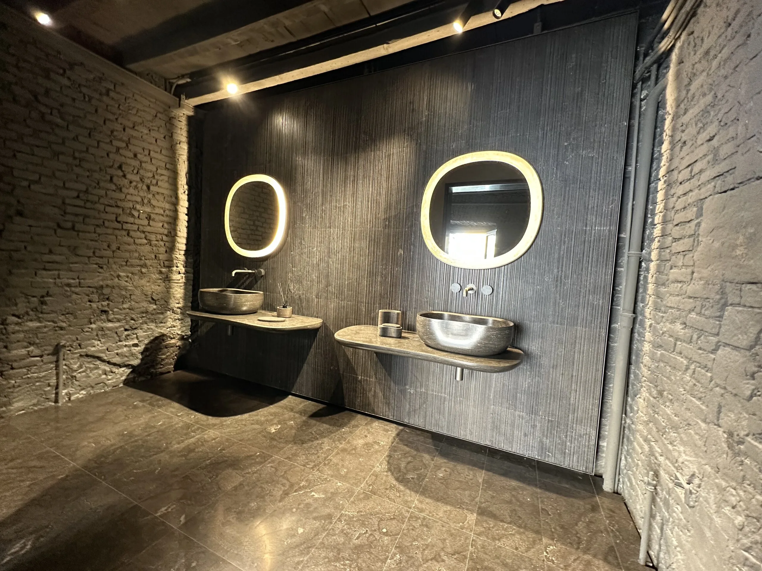 Zweifach-Waschtisch in Natursteinoptik mit beleuchteten Spiegeln vor rustikaler Ziegelwand im Brera Design District Mailand