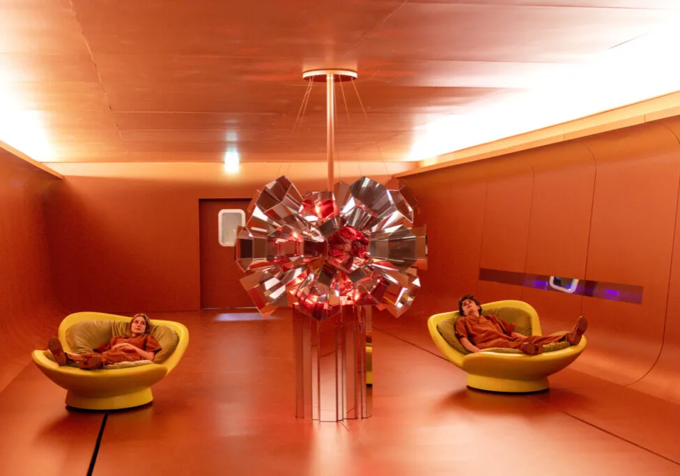 Zwei Personen entspannen in futuristischer, rot getauchter Rauminstallation mit zentralem Lichtobjekt auf dem Salone del Mobile 2025.